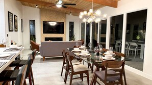 A modern dining area with a large table set for six, surrounded by wooden chairs. A flat-screen TV is mounted on the wall above the seating area, and a ceiling fan is visible.