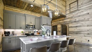 A modern kitchen with grey cabinets, a large island with bar stools, stainless steel appliances, and wooden walls. - Family & Group Cabin w/ Hot Tub & Game Room (Broken Bow)