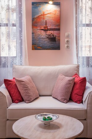 A living room with a beige sofa, red and pink cushions, a round wooden coffee table, and a painting of a sunset over water on the wall.