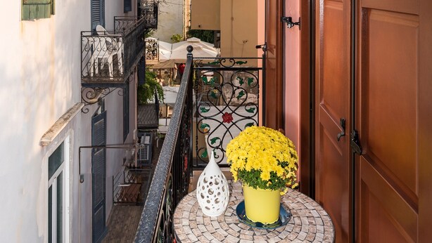 Une rue étroite, avec un clocher en arrière-plan, un balcon agrémenté d’une table et de fleurs, et un bâtiment dont la porte en bois est fermée.