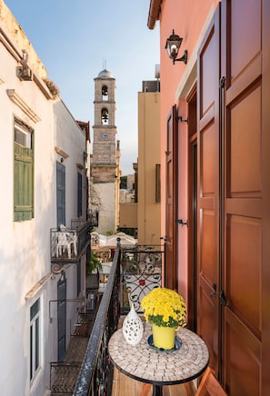 Eine enge Straße mit einem im Hintergrund sichtbaren Glockenturm, einem Balkon mit Tisch und Blumen sowie einem Gebäude mit einer geschlossenen Holztür.