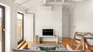 Ein modernes Wohnzimmer mit Fernseher auf einem weißen Konsolentisch, einem Essbereich mit Glastisch und Stühlen sowie einem Balkon mit Geländer.