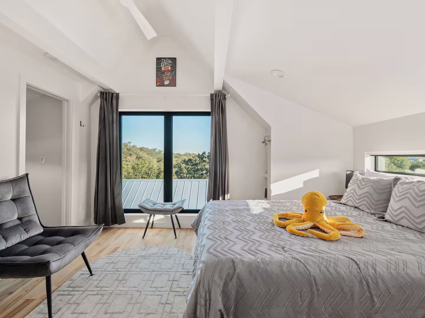 A modern bedroom with a large bed, a chair, a window with curtains, and a view of the outdoors.