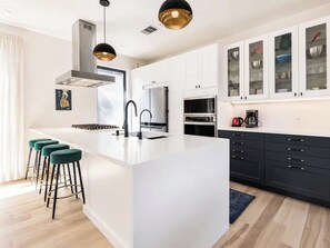 A modern kitchen with a central island, bar stools, a coffee machine, and a view of the living area. - Bright and Clean 7BR East Austin Retreat - Sleeps 12 + Private Yard w/ Fire pit (Austin)
