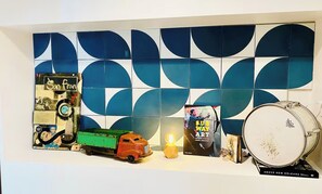 A wall with a geometric tile pattern, a vintage toy truck, a poster, a drum, and a small table with a lamp.