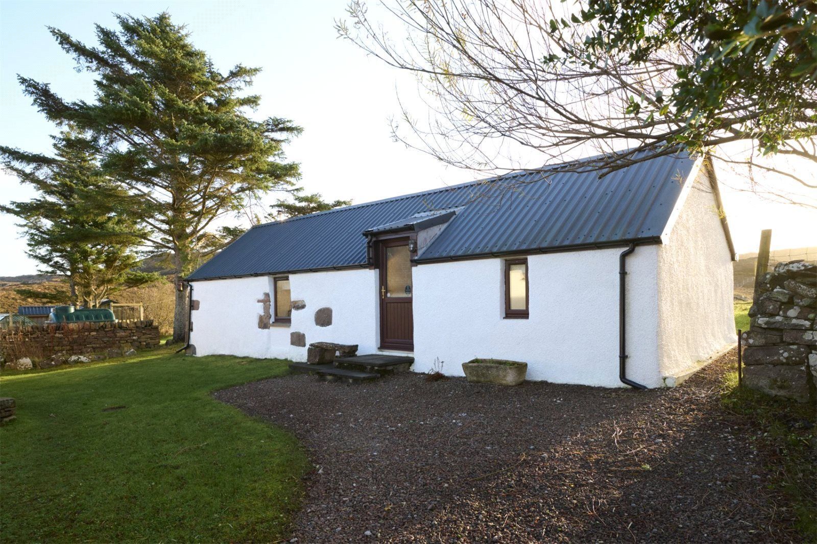 A small, white building with a dark roof, a stone wall, and a gravel driveway.