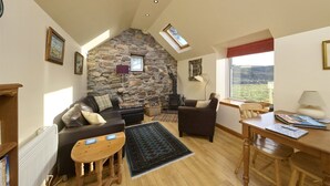 A cozy living room with a stone wall, a fireplace, a sofa, a coffee table, and a view of the outdoors. - The Smithy House & Cottages (Stoer)
