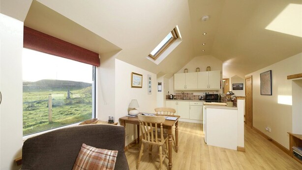 A cozy living room with a view of a grassy landscape, a dining area with a table and chairs, and a kitchen with cabinets and appliances. - The Smithy House & Cottages (Stoer)