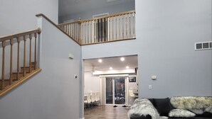 Un vaste espace de vie avec un escalier menant à un deuxième étage, un canapé noir agrémenté d’un plaid en fausse fourrure et un coin repas visible à travers des portes vitrées.