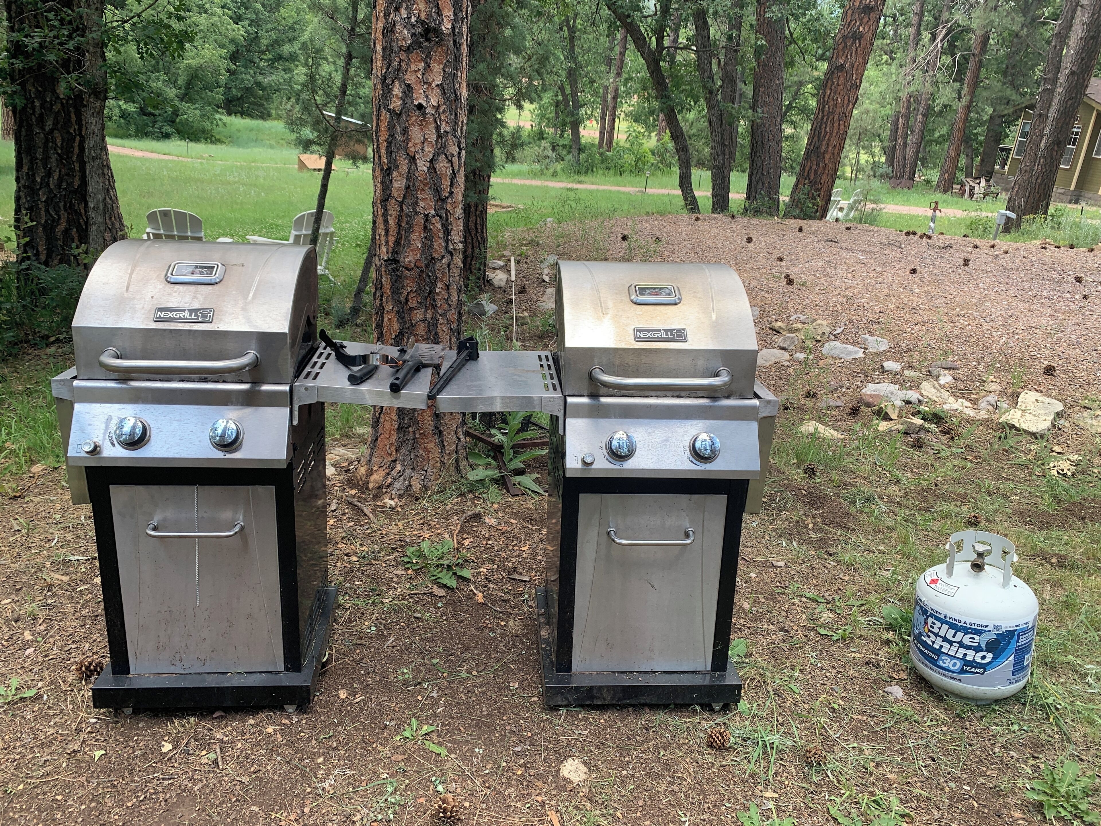 Two NEXGRILL gas grills with a propane tank nearby in a wooded area.