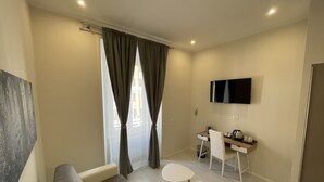 A modern living room with a sofa, two small tables, a desk with a chair, a flat-screen TV, and a window with curtains. - Quintus Luxury Suites (Rome)
