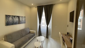 A modern living room with a grey sofa, a small round table, and a wooden desk with a chair. There is a painting on the wall and a window with curtains. - Quintus Luxury Suites (Rome)