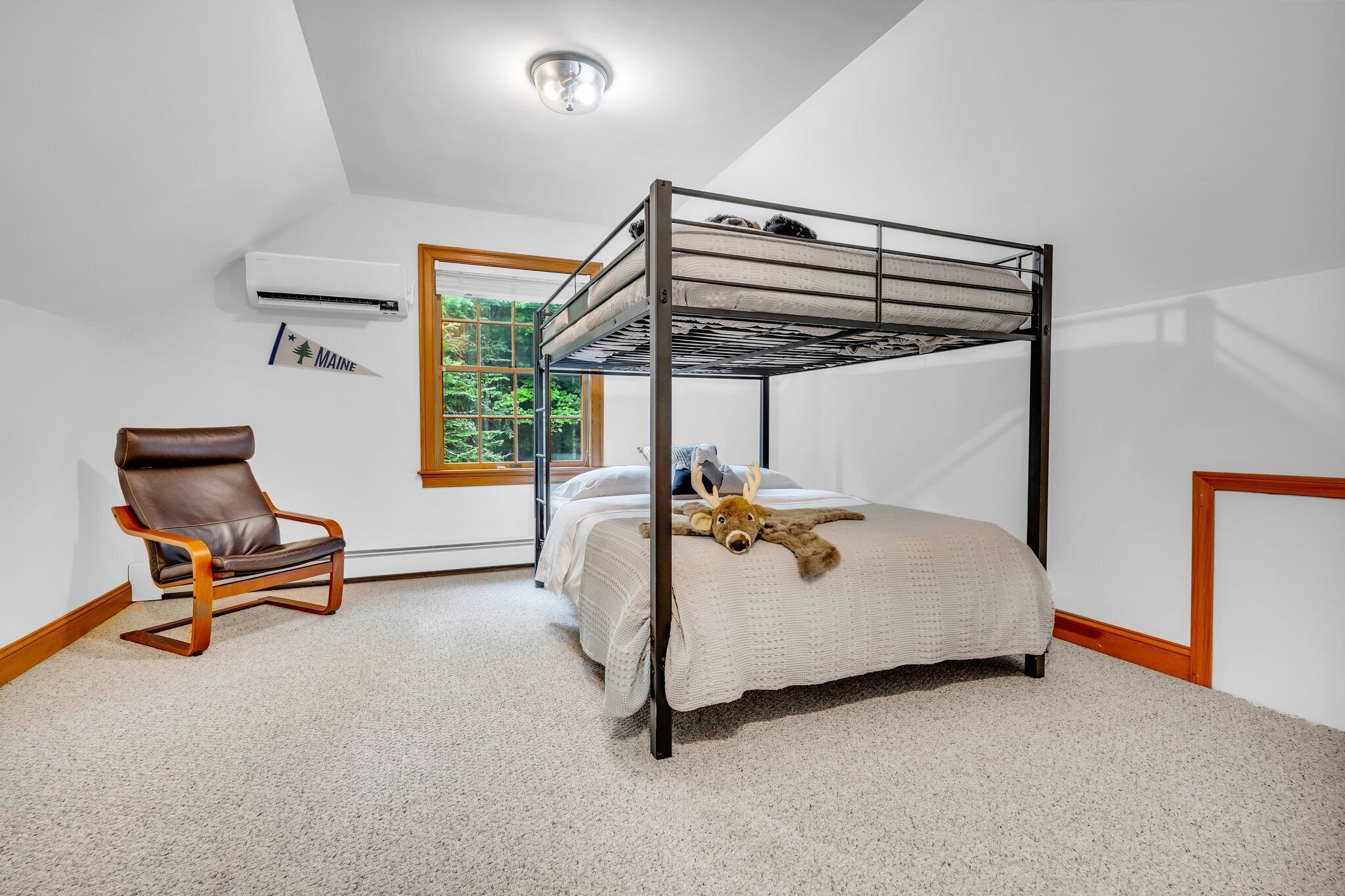 A bunk bed with a headboard, a brown leather chair, a window with a view of greenery, and an air conditioning unit on the wall.