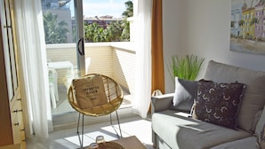 Un salon avec un canapé, un fauteuil en osier et une vue sur les immeubles à travers une porte vitrée.