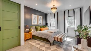 Un dormitorio moderno con una puerta verde, una cama con muchos almohadones, una mesita de noche, un ventanal con cortinas y una araña.