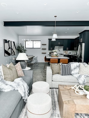 A modern living room with a grey sofa, ottomans, and a coffee table. There is a kitchen area with dark cabinets and a refrigerator. - Luxury Mountain Home with Hot Tub in Angel Fire (Angel Fire)