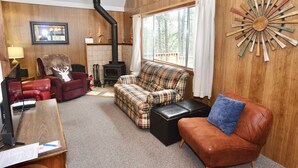 A cozy living room with a fireplace, plaid sofa, leather armchair, and a wooden wall decor piece. - Mint Chip Cabin - Pet friendly - Forested - Large deck - Fire pit (Cascade)