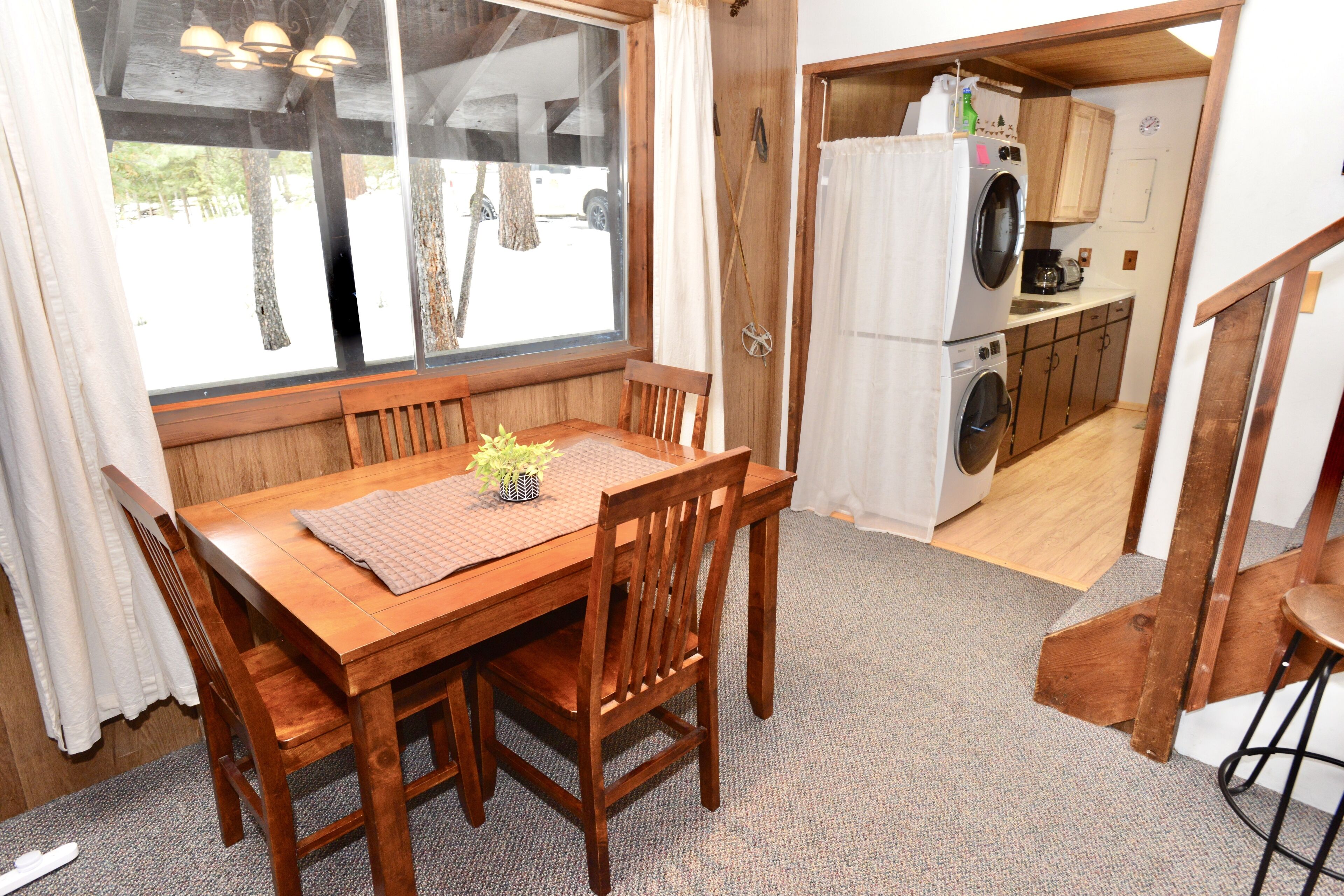 A wooden dining table with chairs, a small plant, and a view of a kitchen with a washing machine.