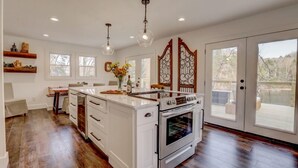 A spacious kitchen with a large island, white cabinetry, and a built-in oven. There are two pendant lights above the island, and a decorative metal partition with a window. - Lakefront Home w/ Dock · King Suite · Stunning Paradise · Voted Best of Kentucky (Laurel County)