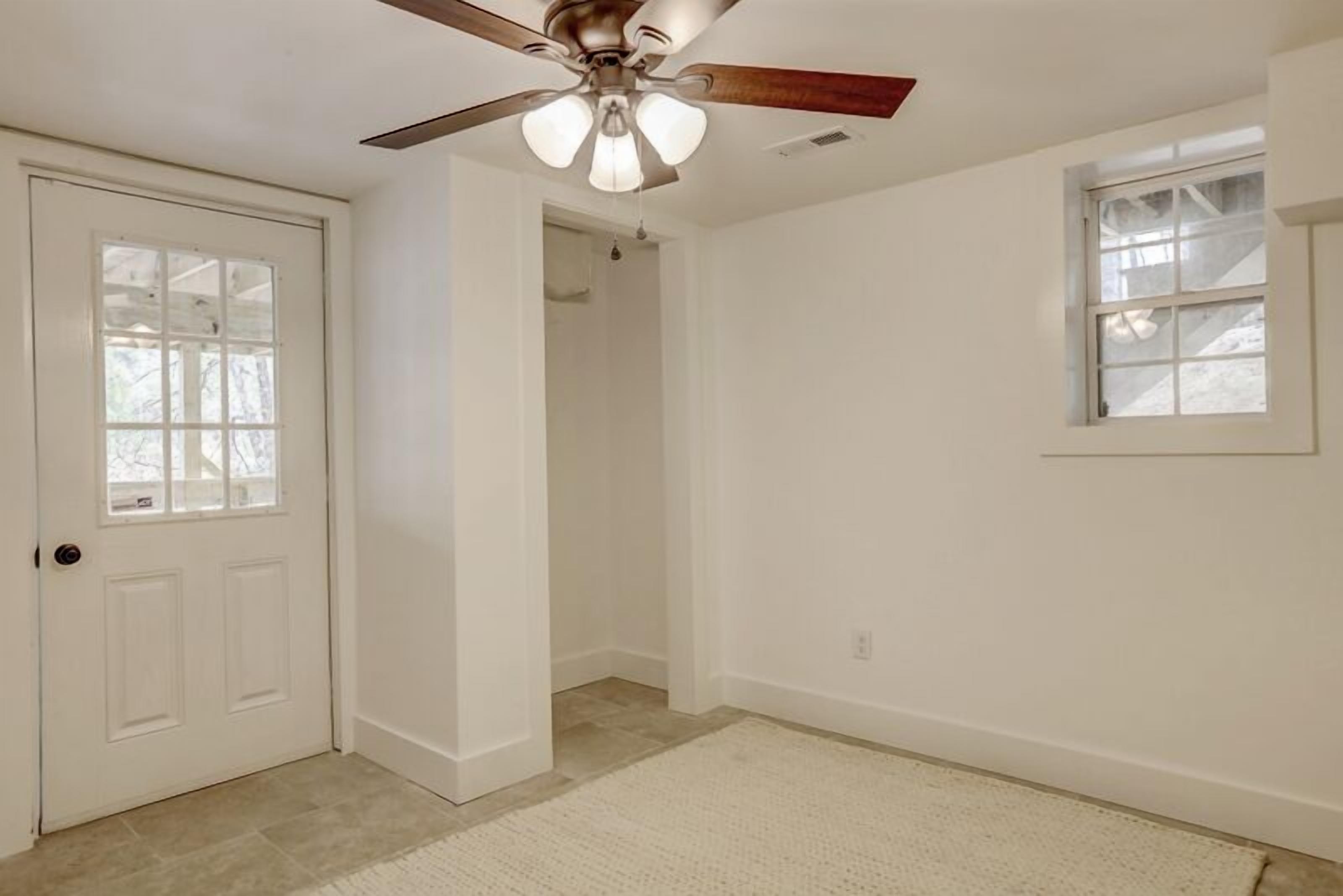 A room with a white door, a ceiling fan, a window, and a door leading to a hallway.
