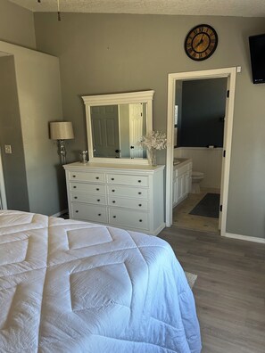 Un dormitorio con una cama grande, un aparador con cajones, un espejo, un reloj y una puerta que da a un baño.