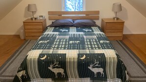 A bedroom with a sloped ceiling, a large window with a view of trees, a bed with a quilt featuring deer and stars, two bedside tables with lamps, and a wooden headboard.