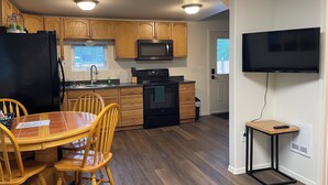 A compact kitchen with wooden cabinets, a black refrigerator, a microwave, and a stove. There is a dining table with chairs and a television mounted on the wall.