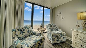 Een kamer met een groot raam met uitzicht op het strand, twee gestreepte banken, een tafeltje en een dressoir met decoratieve spullen.