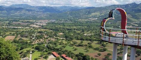 Un mirador pintoresco con barandilla metálica, una estructura roja y blanca, y una vista de un valle con edificios y vegetación.