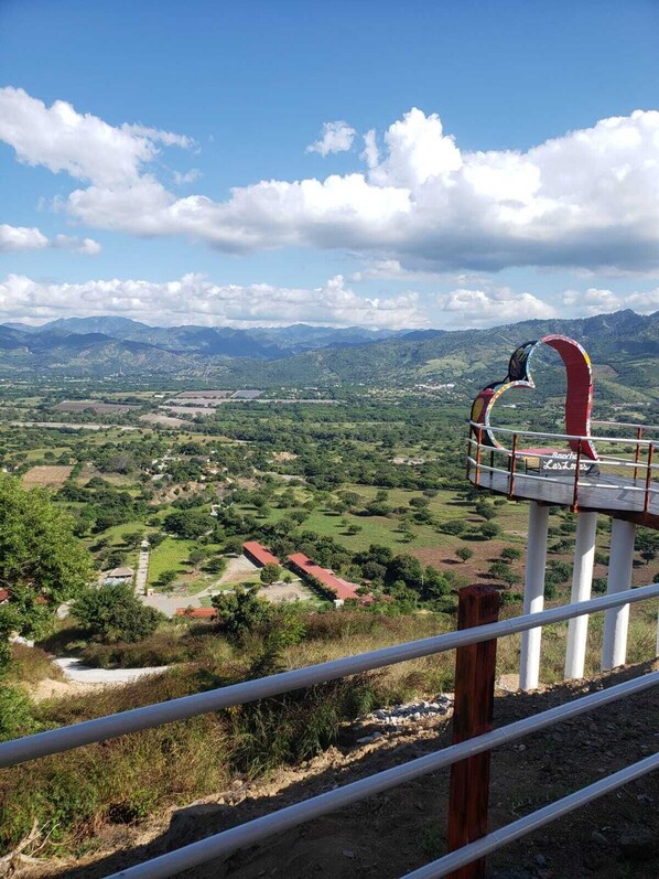 Un mirador pintoresco con barandilla metálica, una estructura roja y blanca, y una vista de un valle con edificios y vegetación.