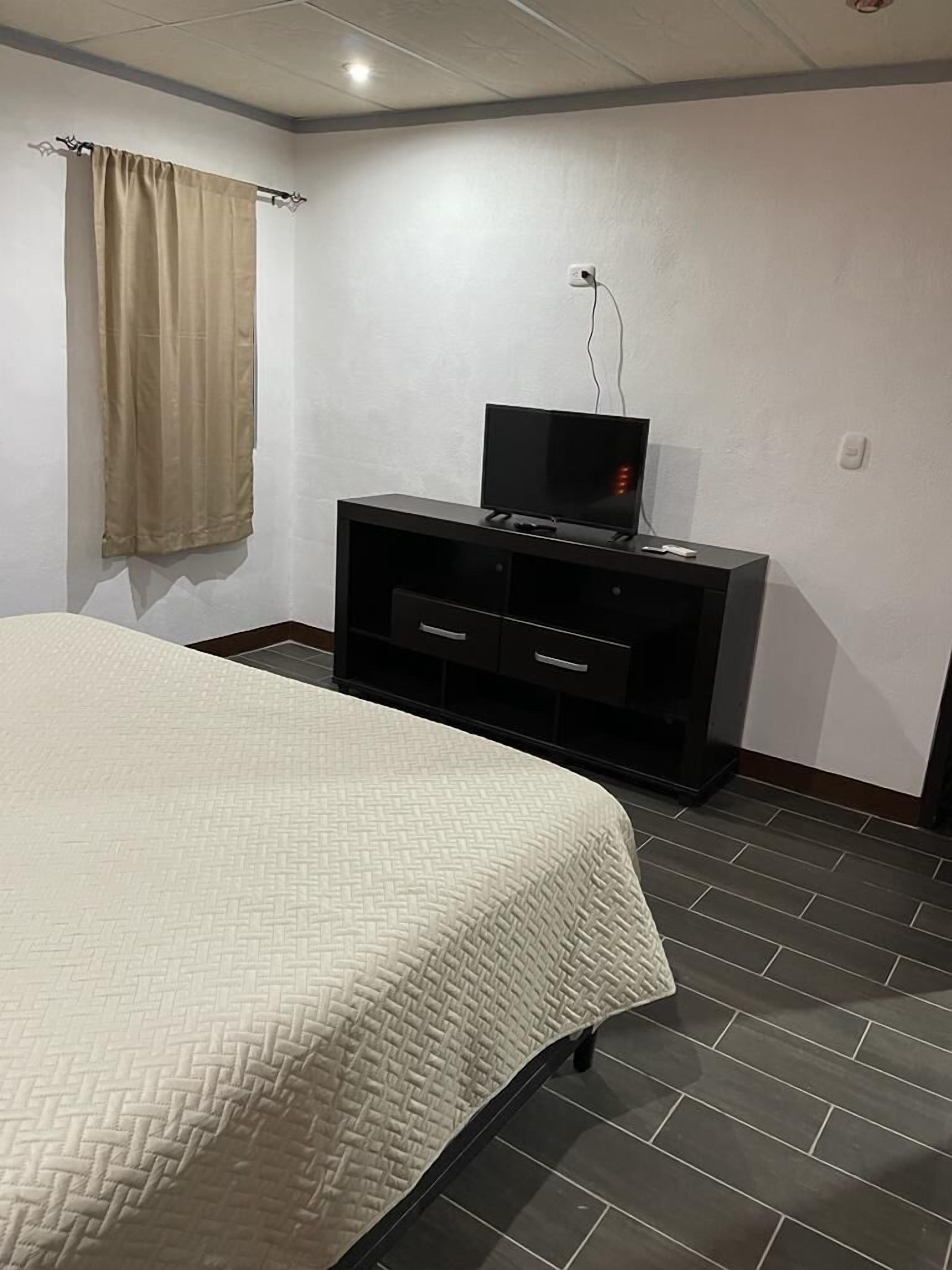 A bedroom with a bed, a TV on a black stand, a curtain, and a wall socket.