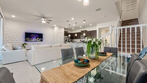 A modern living room with a glass dining table, white sofas, and a kitchen in the background.