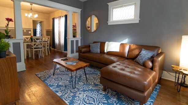 A living room with a brown leather sofa, a wooden coffee table, and a patterned rug. There is a dining area visible in the background.