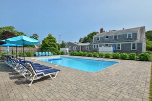 A rectangular swimming pool with lounge chairs and umbrellas, surrounded by a brick patio and a two-story building with a red door.