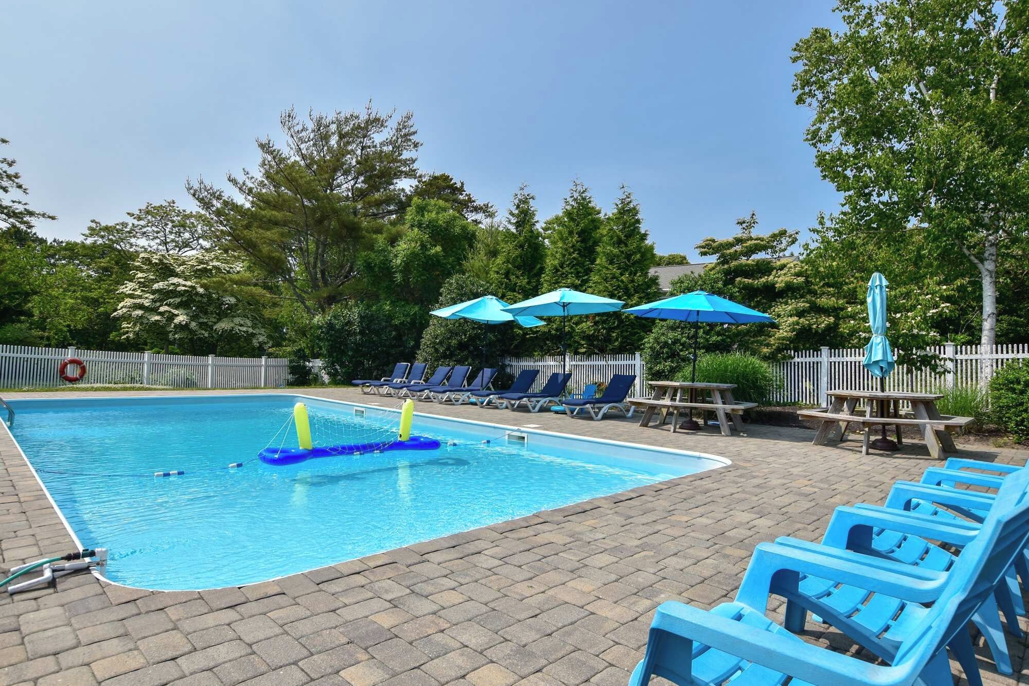 A swimming pool with blue lounge chairs and umbrellas, surrounded by trees and a white fence.