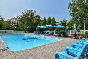 A swimming pool with blue lounge chairs and umbrellas, surrounded by trees and a white fence. - Heated Pool AC 1/2 Mile to Beach! (Harwich)