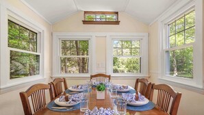 Uma sala de jantar com mesa de madeira posta para seis pessoas, rodeada por cadeiras, janelas amplas com vista para árvores e um pequeno claraboia acima.