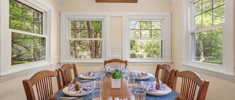A dining room with a wooden table set for six, surrounded by chairs, large windows with a view of trees, and a small skylight above.