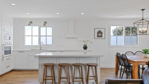 Eine moderne Küche mit weißen Schränken, einer zentralen Kochinsel mit Hockern, einem Essbereich mit Holztisch und Stühlen sowie großen Fenstern für natürliches Licht.