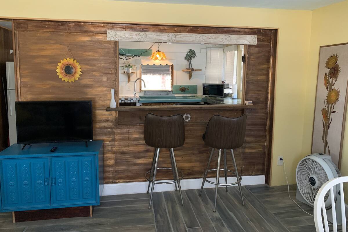 A kitchen area with a wooden counter, two bar stools, a sink, a microwave, and a TV on a blue cabinet.