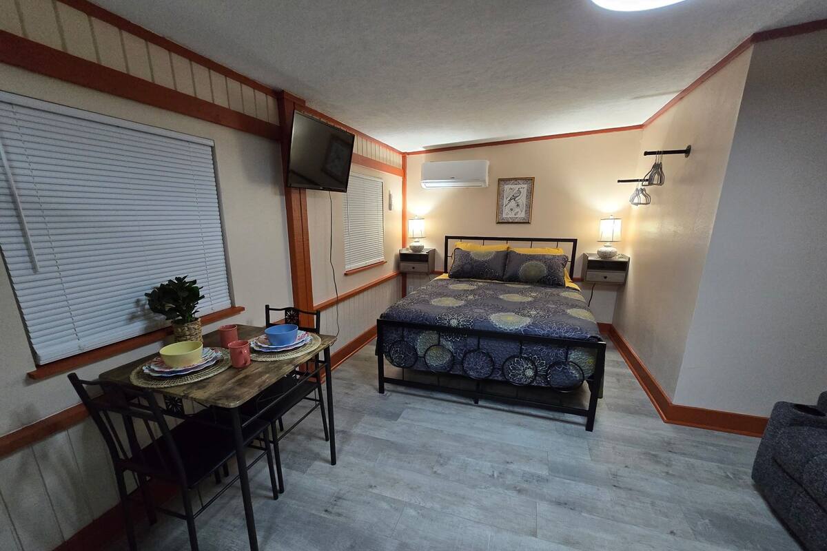 A compact bedroom with a bed, bedside tables, a TV, and a dining table with chairs.