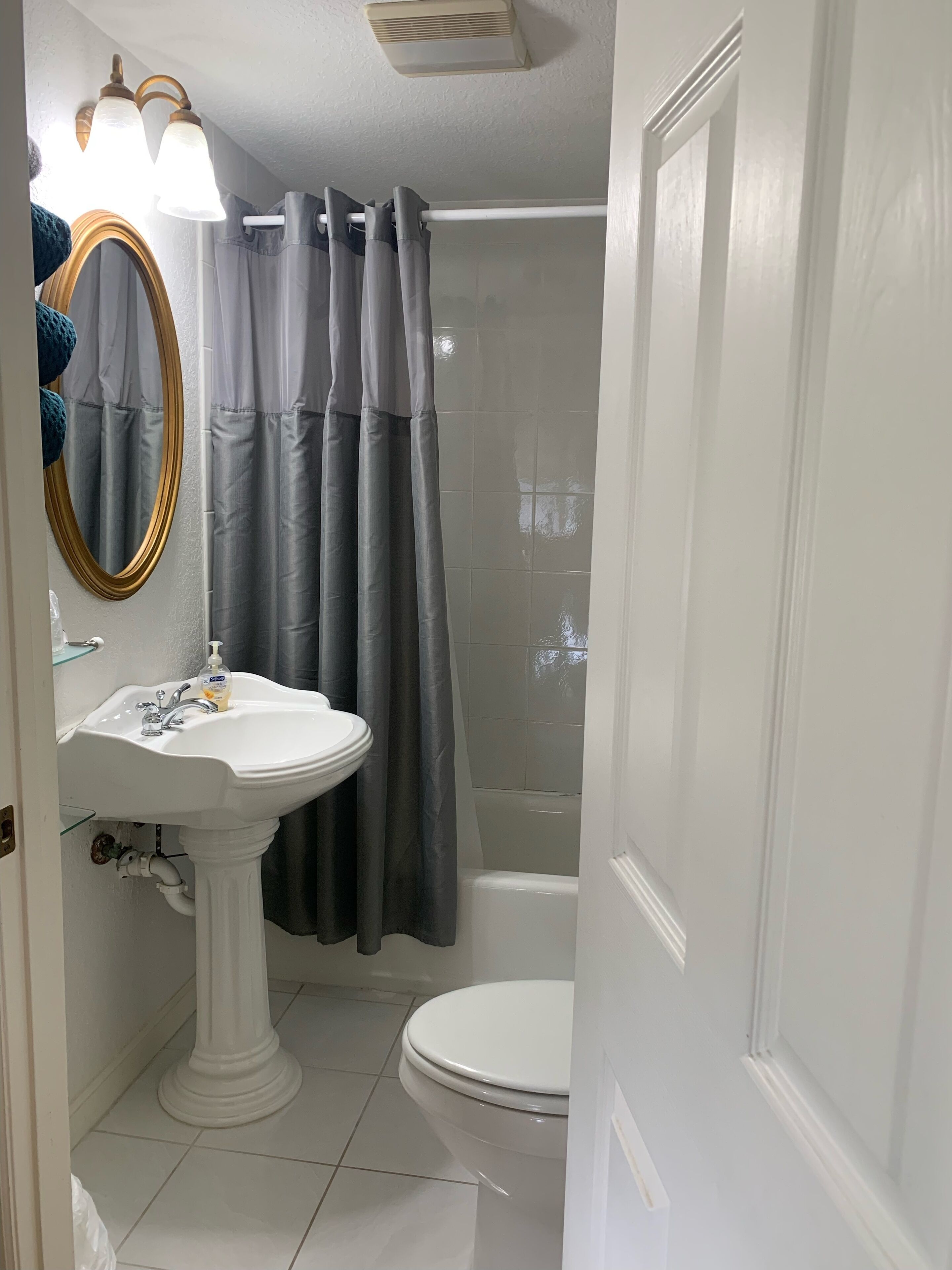 Un baño con lavamanos pedestal, inodoro, ducha con cortina y espejo con marco dorado.