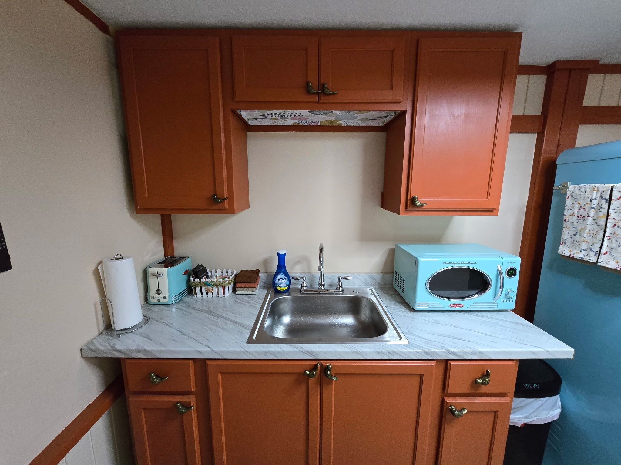 A compact kitchen with wooden cabinets, a sink, a toaster, a microwave, and a blue appliance.