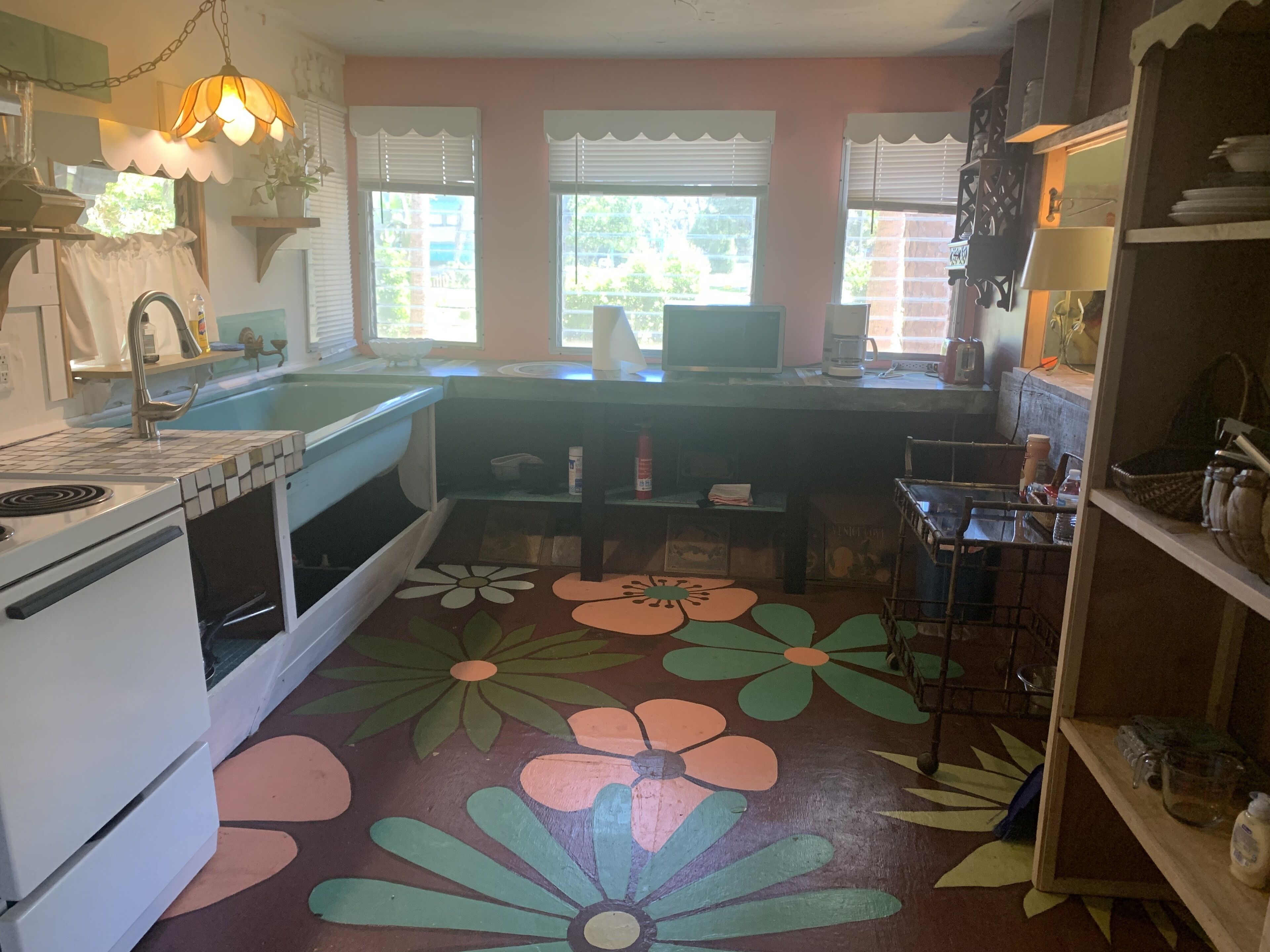Une cuisine équipée d’une cuisinière, d’un évier et d’un sol décoré de motifs floraux colorés. On y trouve des fenêtres avec des stores, un réfrigérateur et une étagère contenant des ustensiles de cuisine.