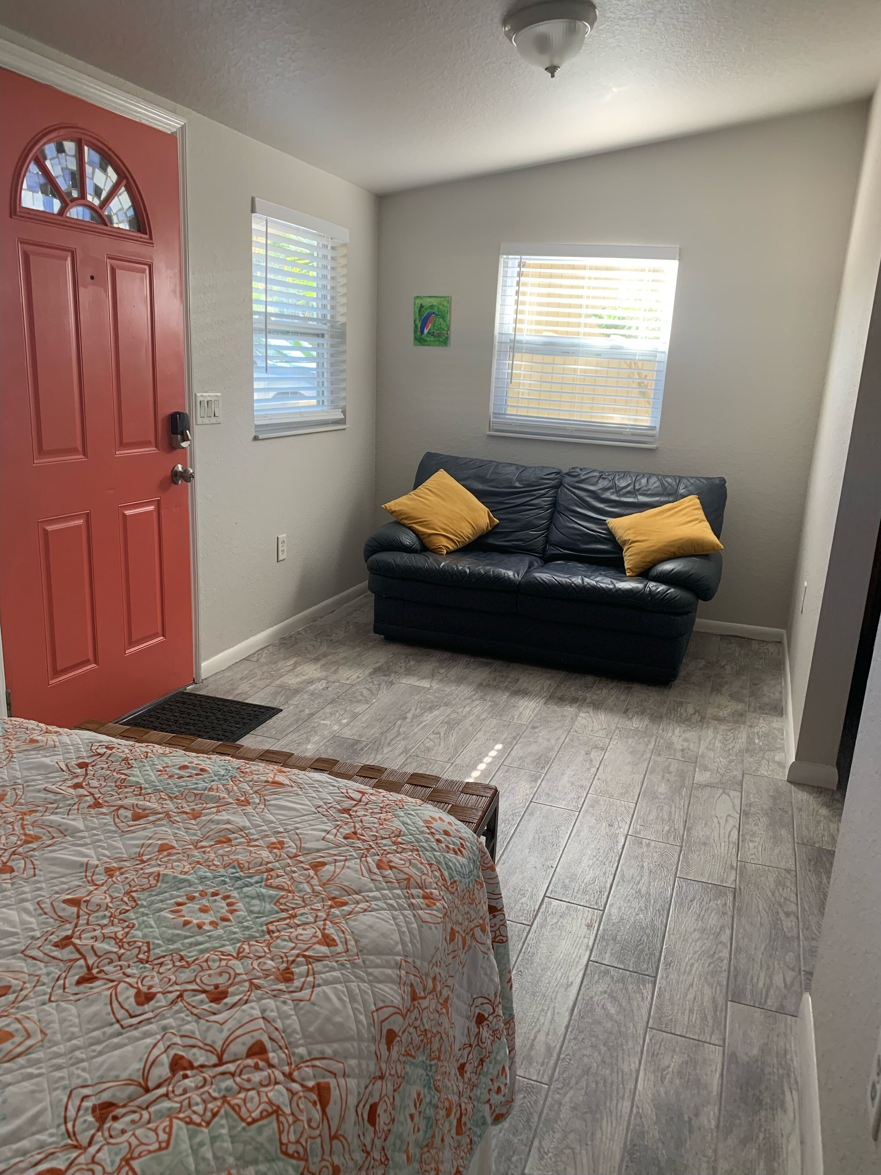 A bedroom with a red door, a black leather sofa with yellow cushions, a patterned bedspread, and a window with blinds.