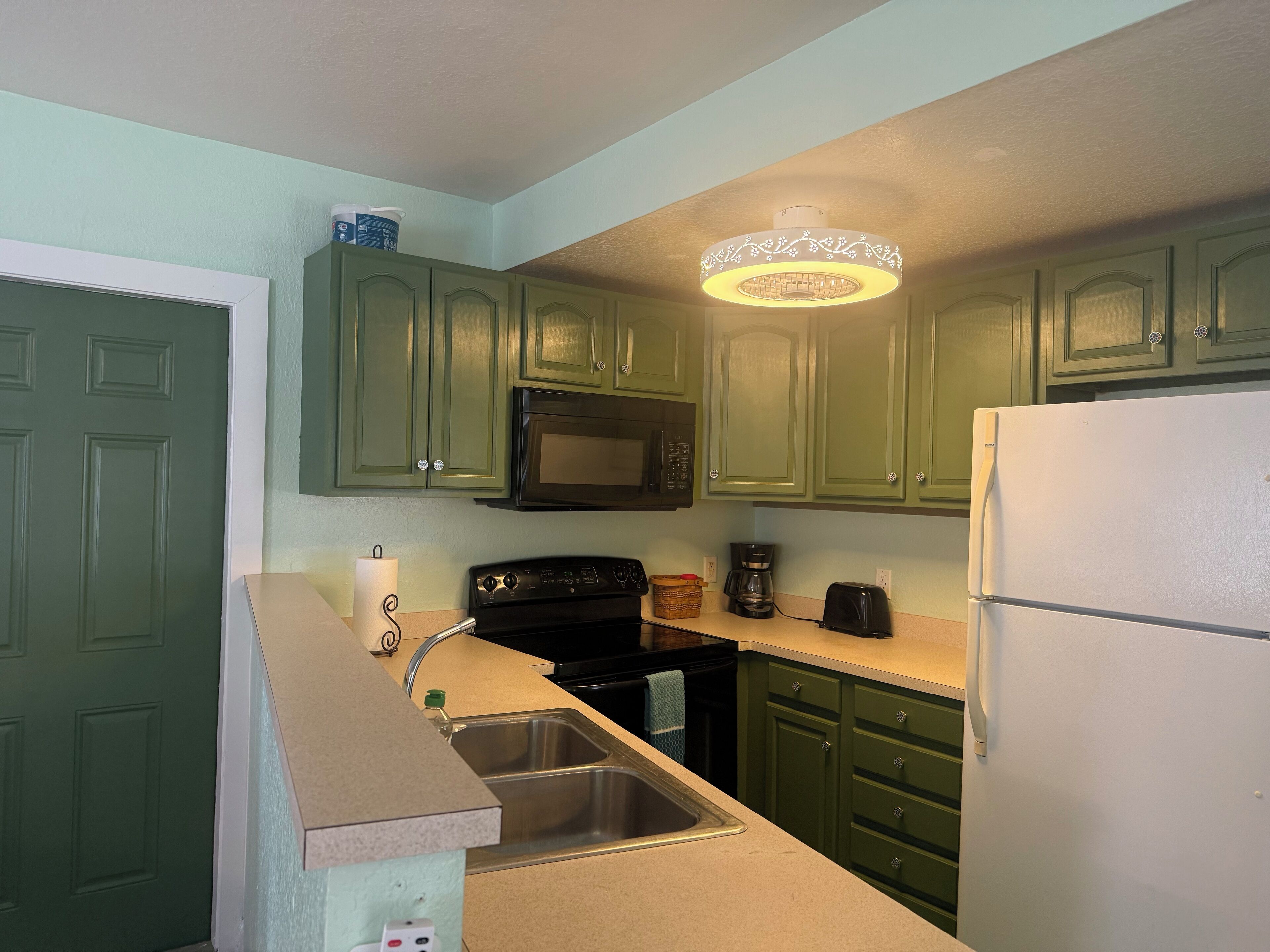 Una cocina con gabinetes verdes, microondas, estufa, fregadero y refrigerador.