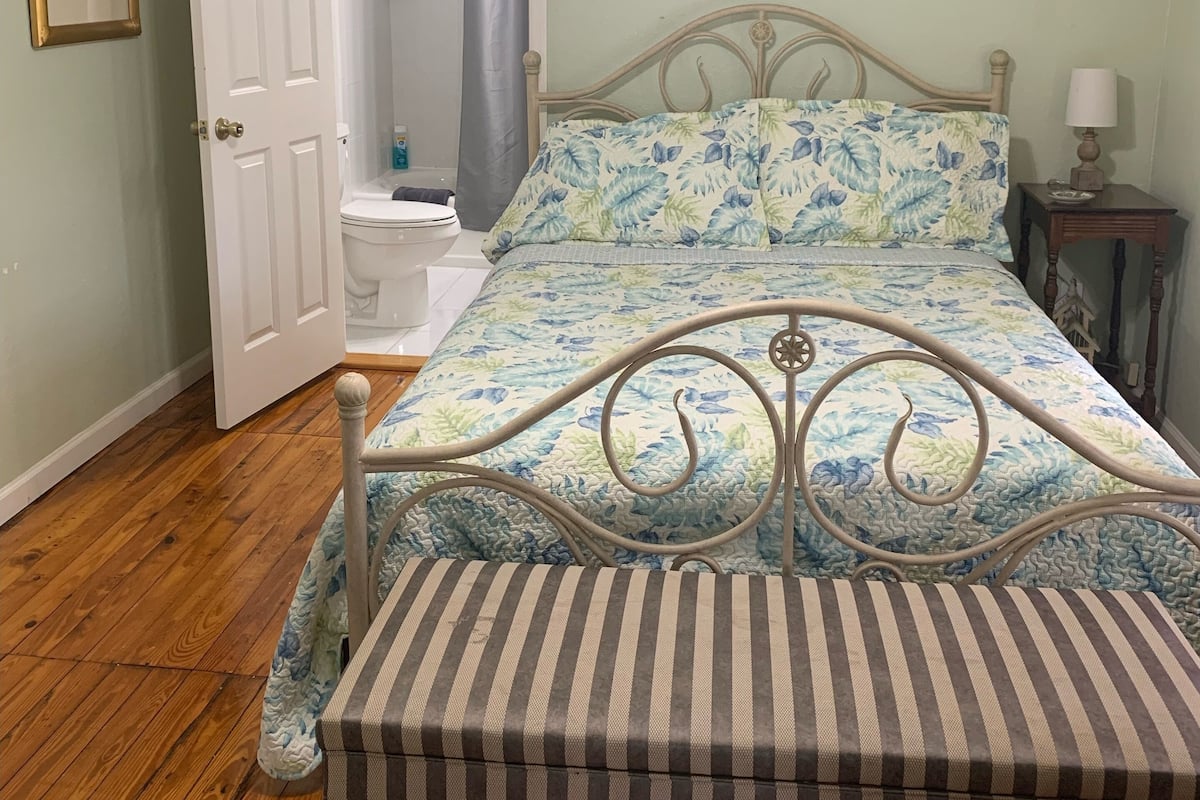 A bedroom with a bed, a bedside table, a lamp, a ceiling fan, a door, and a bathroom visible through an open door.