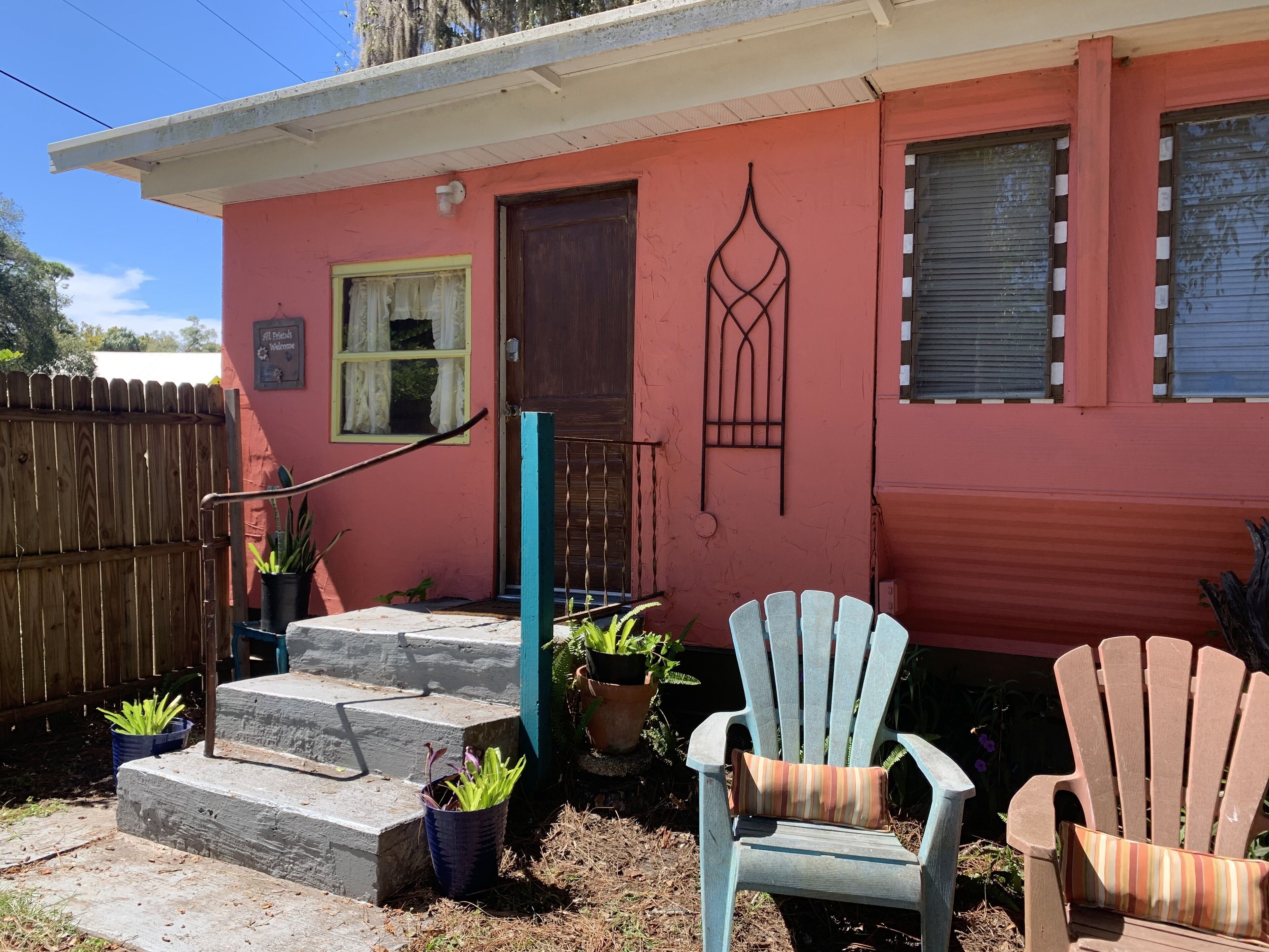 A house with a red exterior, a wooden door, a window with a curtain, and a porch with two Adirondack chairs and potted plants.