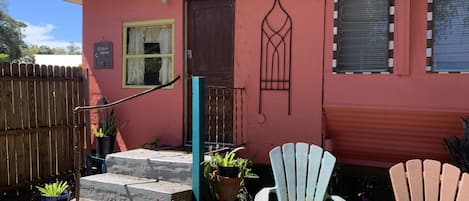 A house with a red exterior, a wooden door, a window with a curtain, and a porch with two Adirondack chairs and potted plants.