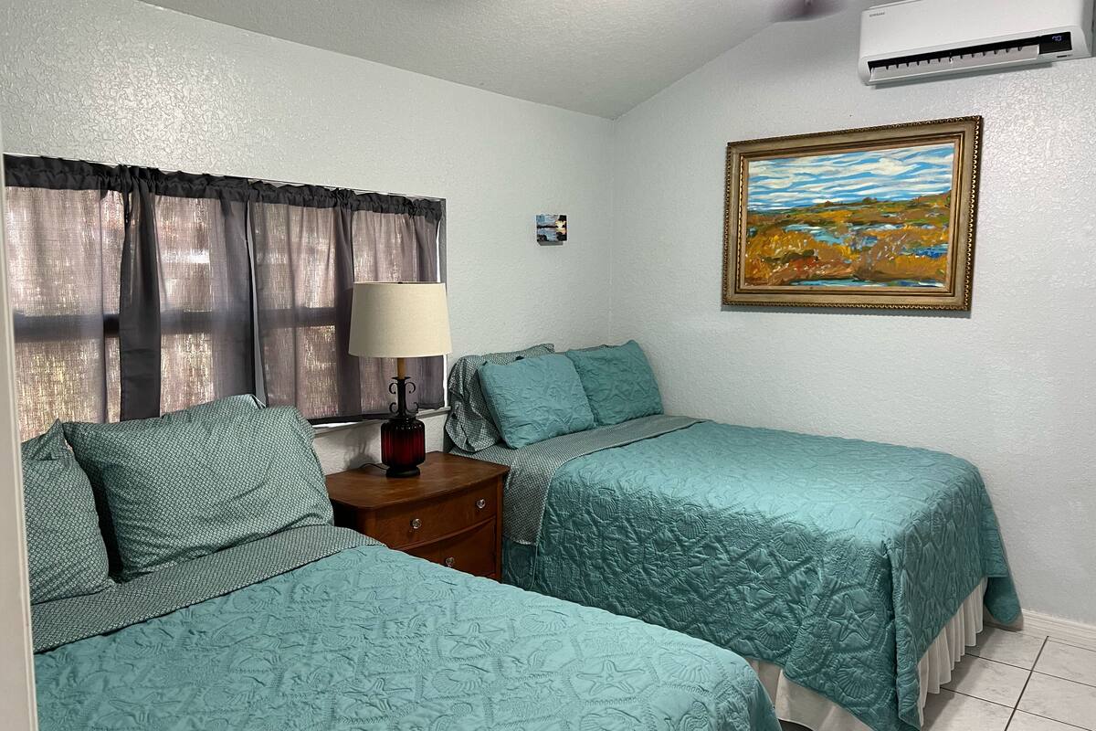 A room with two beds, a nightstand, a lamp, a painting, and an air conditioner.